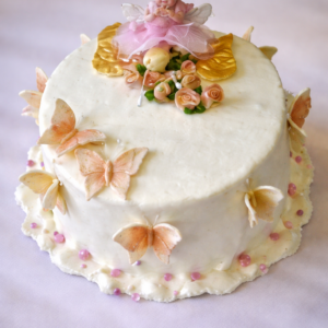 Detská syrová torta s jemným bielym povrchom, ozdobená syrovými ružami, mašličkami a motýľmi, s figúrkou víly v ružových šatách na vrchu.
